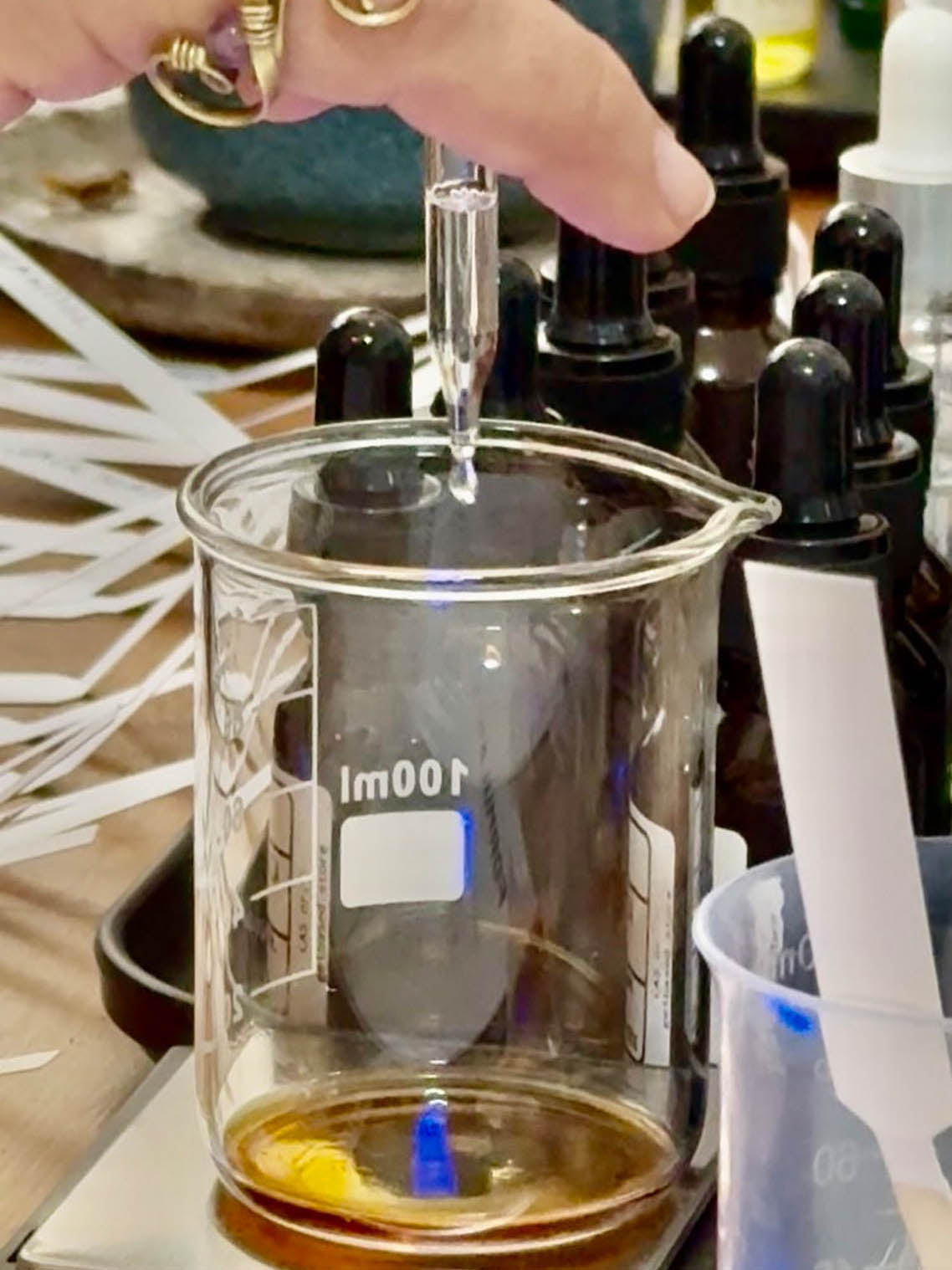 A hand holding a glass dropper above a 100ml glass beaker containing amber liquid, with multiple dark dropper bottles in the background.