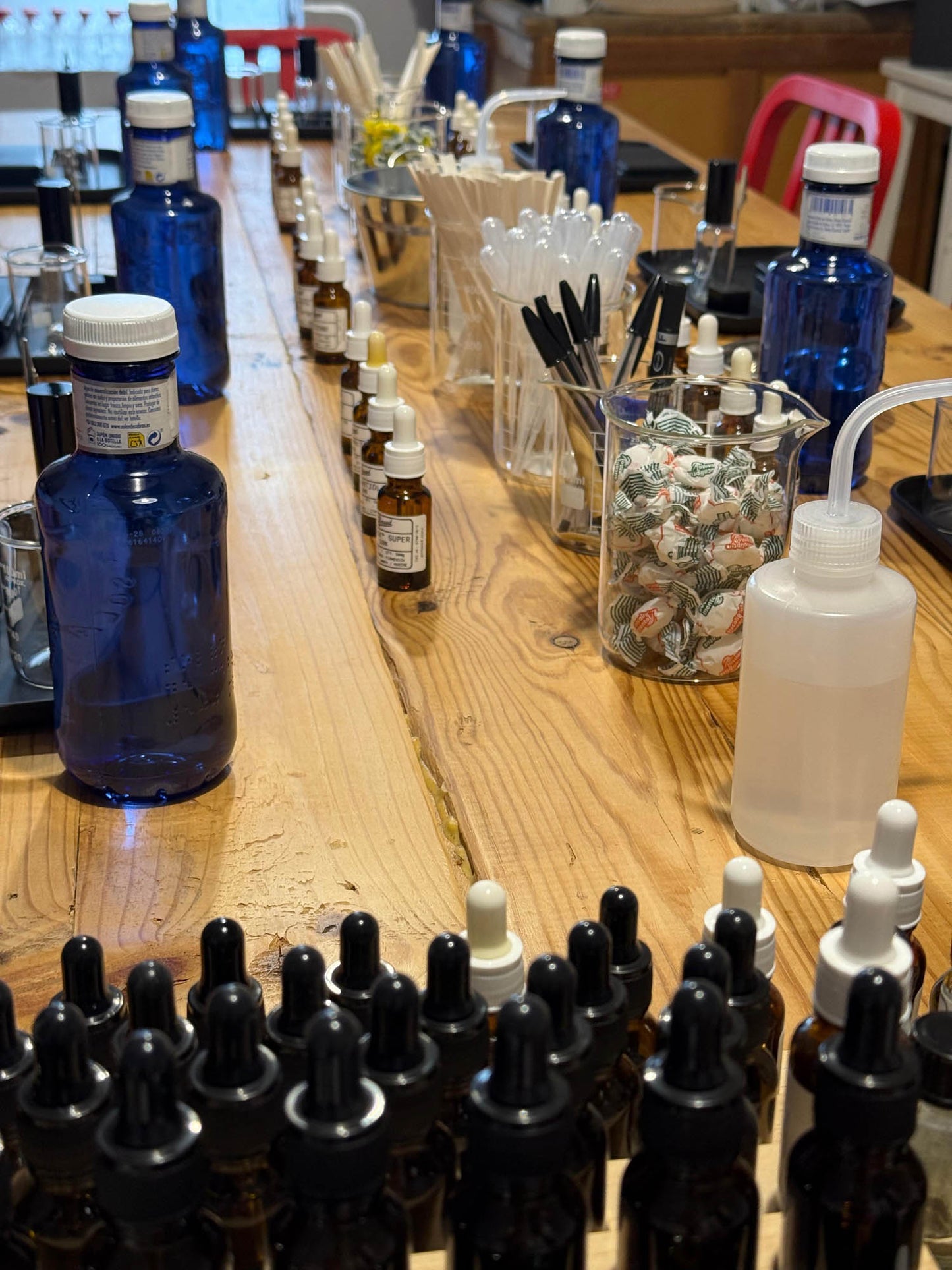 A wooden table filled with small amber glass dropper bottles, several cobalt blue bottles with white caps, a white wash bottle with a clear tube, and containers of mixing tools in a workshop or craft space.