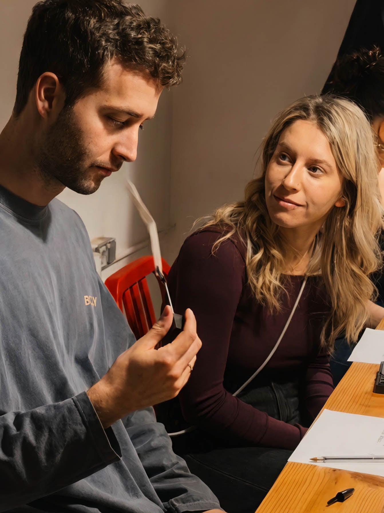 A man in a gray long-sleeve shirt holding a smartphone while a woman in a dark plum top looks on, seated at a wooden table with papers and a pen in a casual indoor setting.