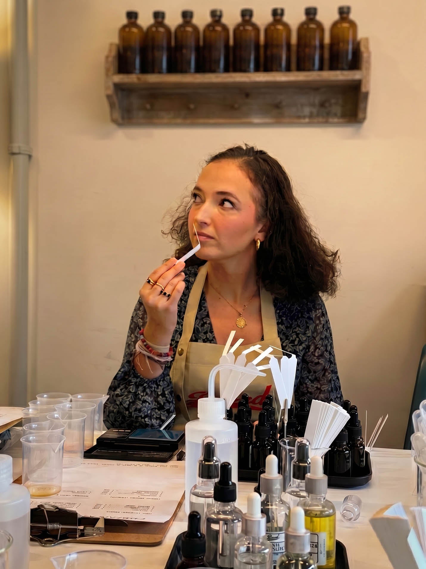 A woman wearing a beige apron over a dark patterned blouse applies lip product while seated at a table filled with cosmetic bottles, droppers, and mixing tools. A wooden shelf with amber bottles hangs on the wall behind her.