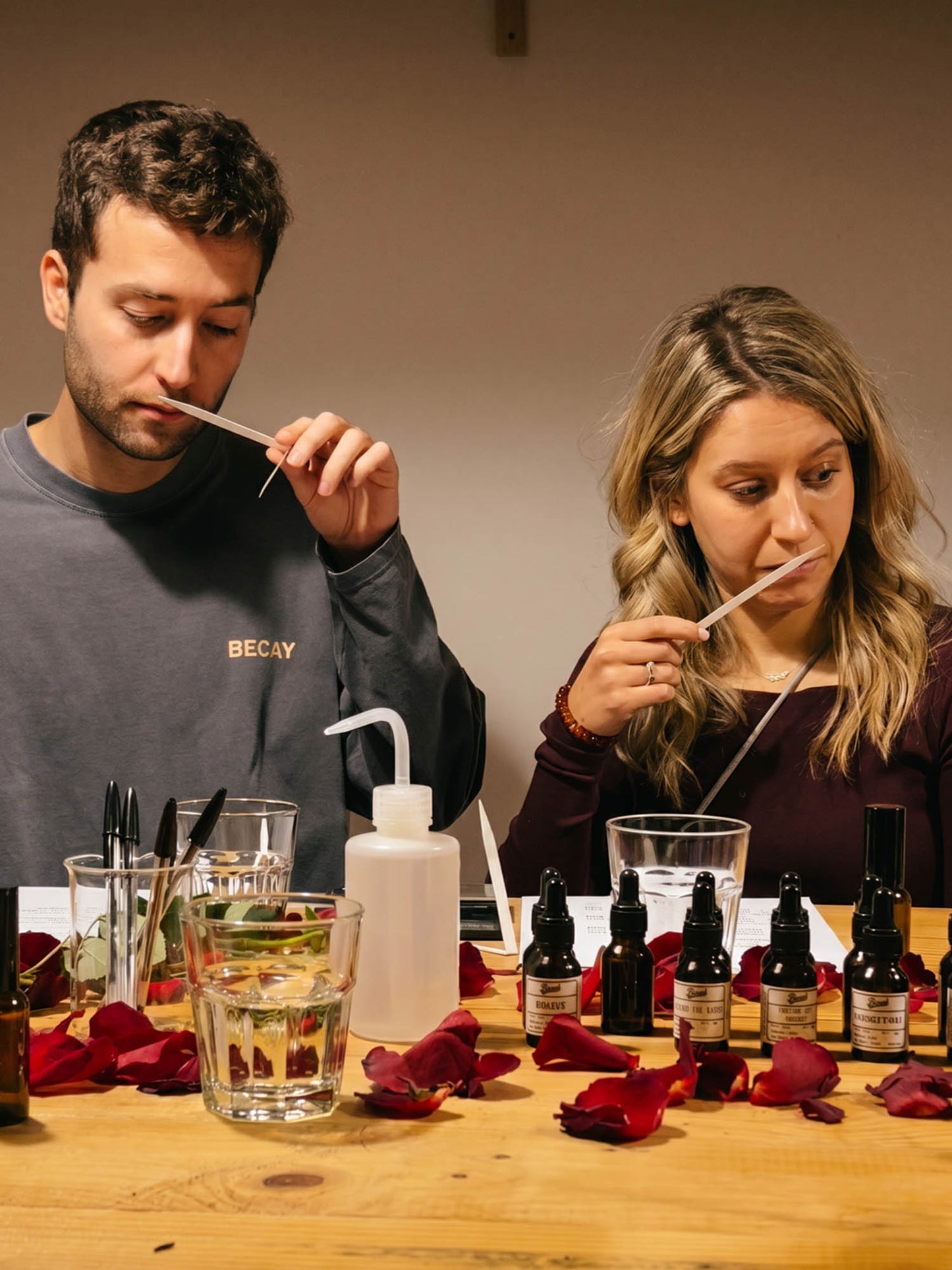 A man and a woman sit at a wooden table, each holding a glass pipette to a small dropper bottle. The table is scattered with red rose petals and contains several small dark bottles, a white squeeze bottle, and glassware.