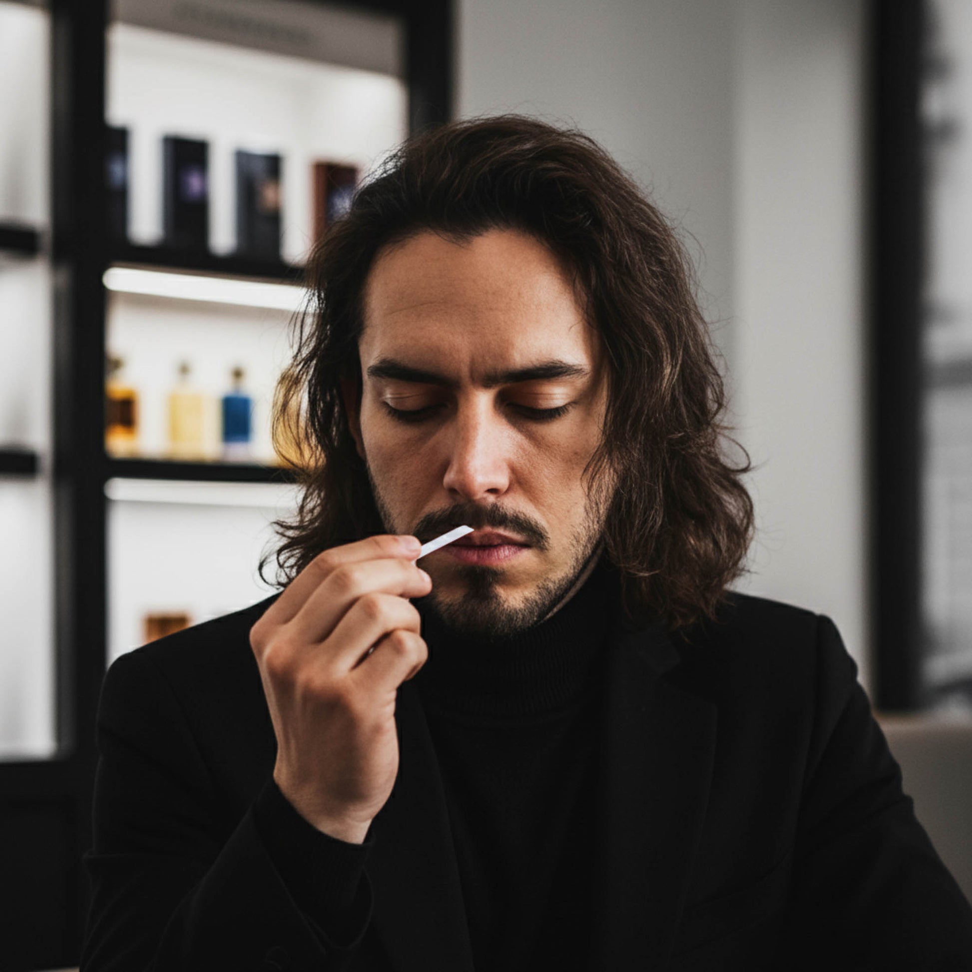A man with long dark hair and a beard, wearing a black turtleneck and blazer, holding a small white object near his mouth in a dimly lit indoor setting with shelves in the background.