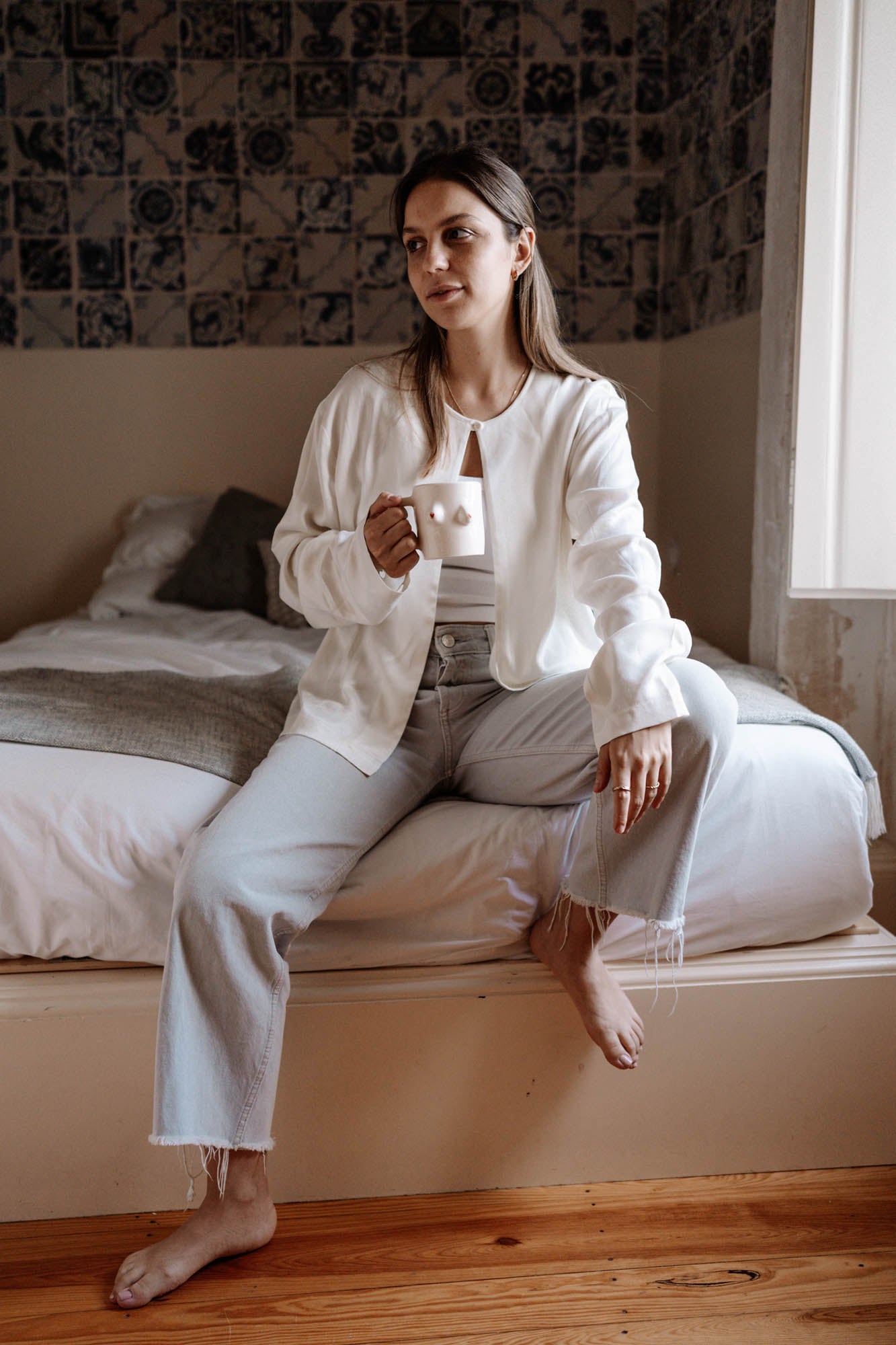 Woman sitting on a bed holding a boobs or breast mug in a cozy bedroom.